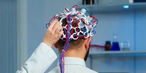 Back view of man patient wearing performant brainwave scanning headset sitting in neurological research laboratory while medical researcher adjusting it, examining nervous system typing on tablet.
